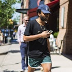 Donald Glover, with a blue cap, a black Guess T-shirt, and green and white shorts, walking down a st...