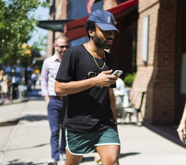 Donald Glover, with a blue cap, a black Guess T-shirt, and green and white shorts, walking down a st...
