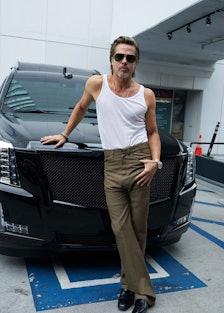 Brad Pitt wearing a white tank top and sunglasses, posing with his hand atop the front of a car