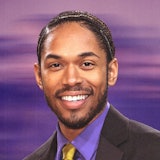 Kelvin Harrison Jr. in a brown suit and purple button down with a gold tie, on a purple background
