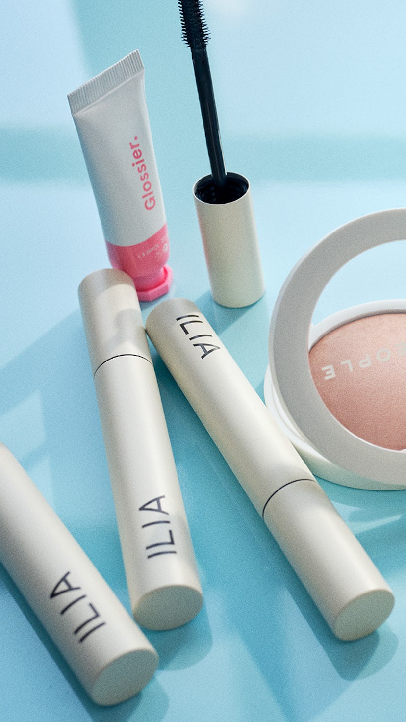 Pregnancy make up products from Ilia, Glossier and Well People placed on a blue table in front of a ...