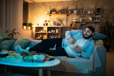 An overweight man sits on the couch, watching TV and eating popcorn.