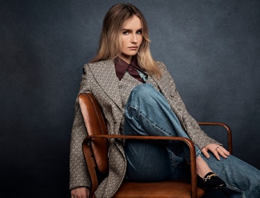 Olivia DeJonge sitting on a brown leather chair with one leg up, wearing coat, vest, silk shirt, tro...