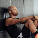 A man playing basketball takes a break by sitting against a padded wall.
