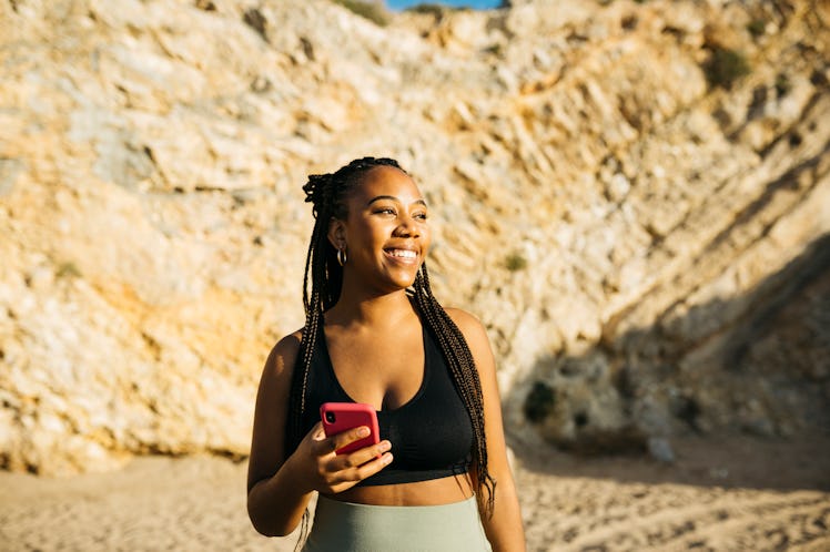 Young woman smiling while holding her cell phone after reading the July 4, 2022 weekly horoscope for...