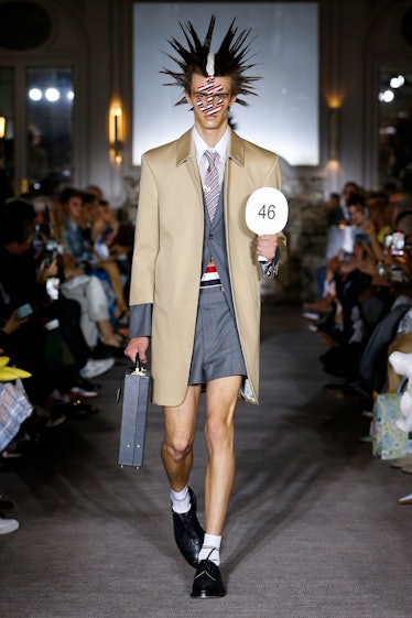 A model wearing a Thom Browne outfit with spiked hair, a beige coat, a gray vest and shorts and a gr...