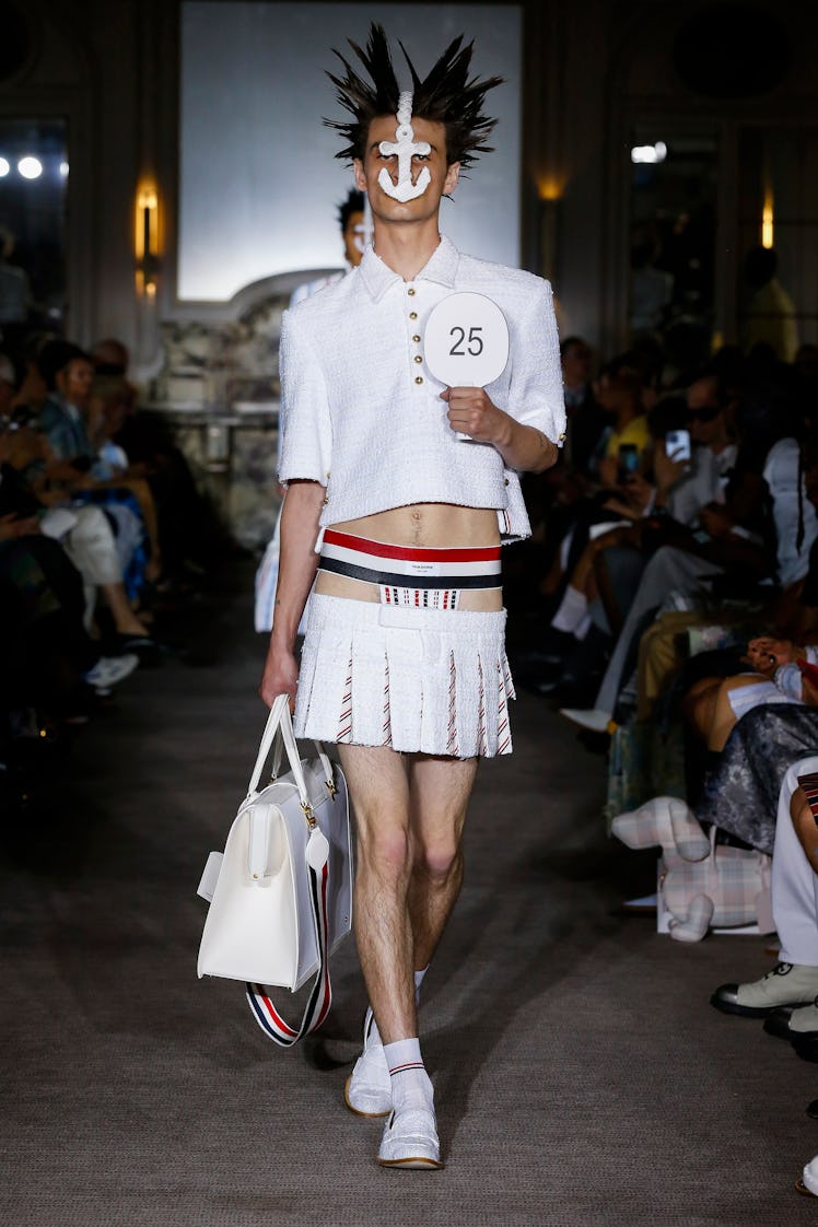 A model wearing a Thom Browne outfit with a white cropped shirt, a white skirt and bag, with spiked ...