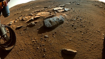 The tire of a Mars rover is visible next to a gray streaked rock on reddish sand.