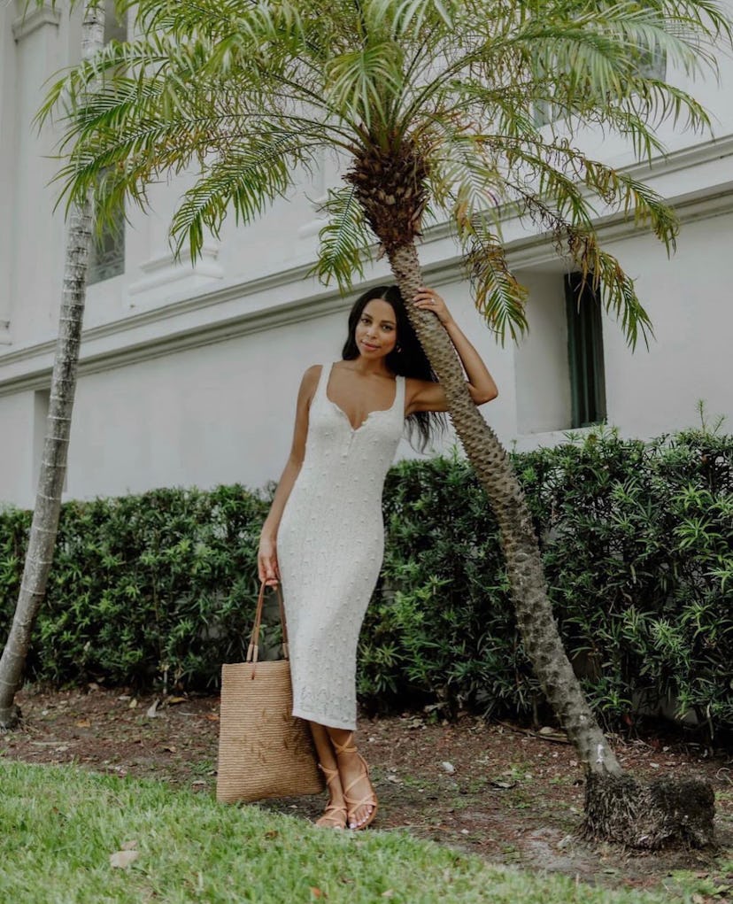 A woman in a beige bodycon dress, holding a wicker basket bag while posing beneath the palm tree
