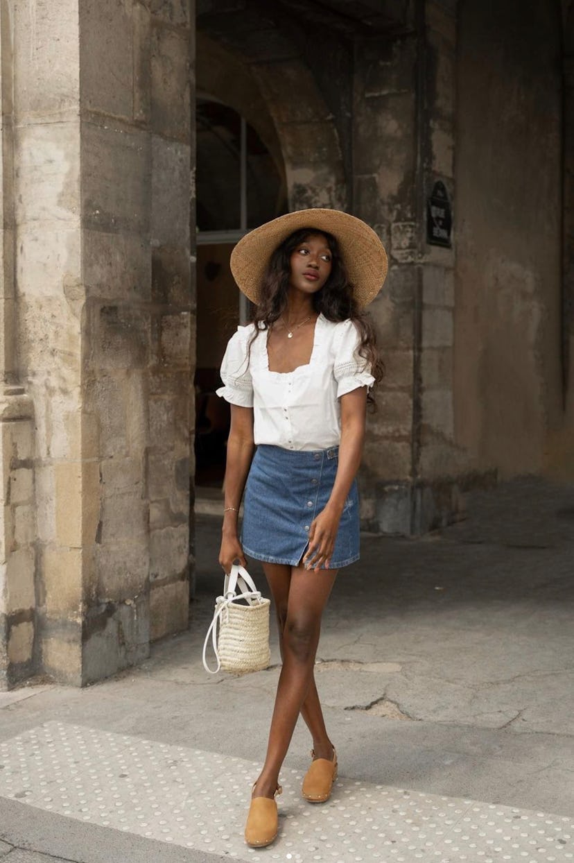 Emmanuelle wearing a mini denim skirt, a white top with puff sleeves, a wicker hat and a matching ba...