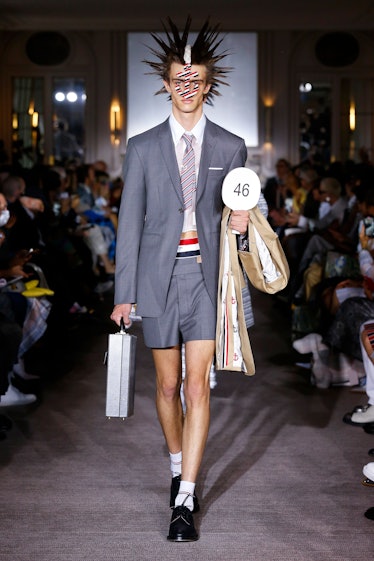 A model wearing a Thom Browne outfit with spiked hair, a gray vest and shorts and a gray briefcase