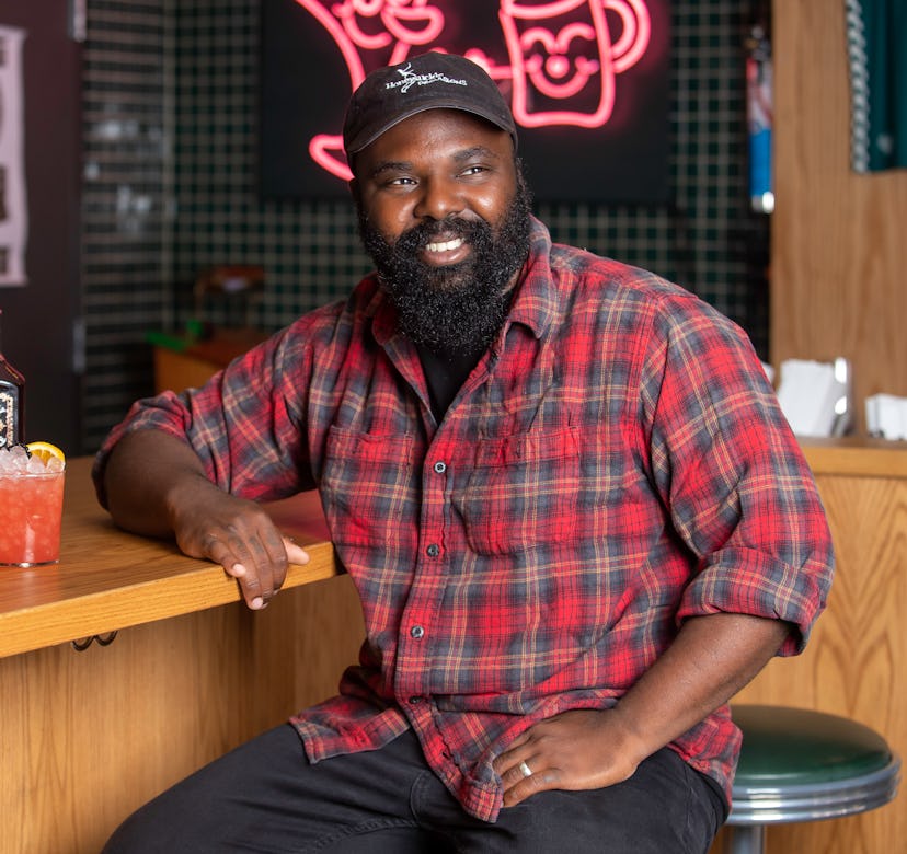 Chef Omar Tate smiling in a red checked shirt, black cap and black jeans while leaning onto a wooden...