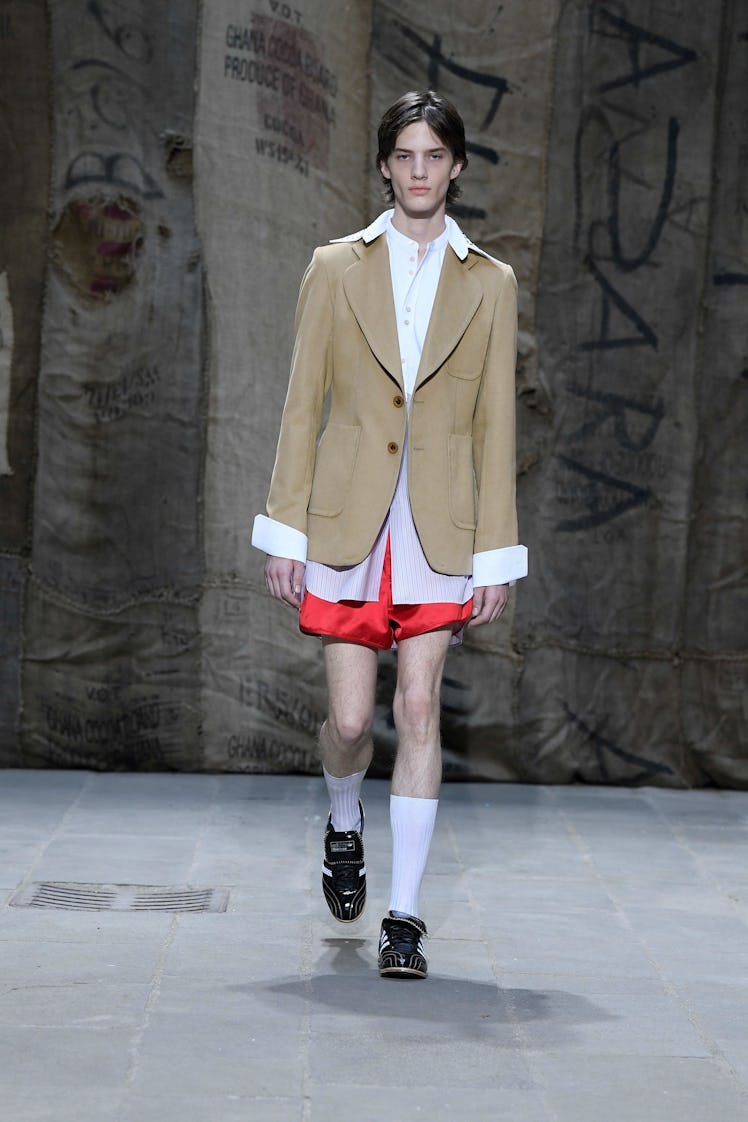 A model wearing a Wales Bonner outfit, with a white button down shirt, a beige blazer and red shorts