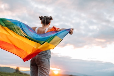 A teen faces away, holding a rainbow flag behind their back, standing outside.