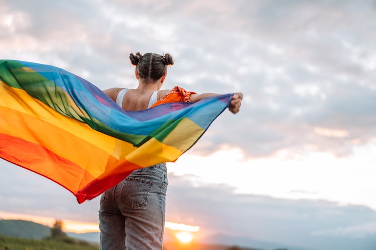 A teen faces away, holding a rainbow flag behind their back, standing outside.