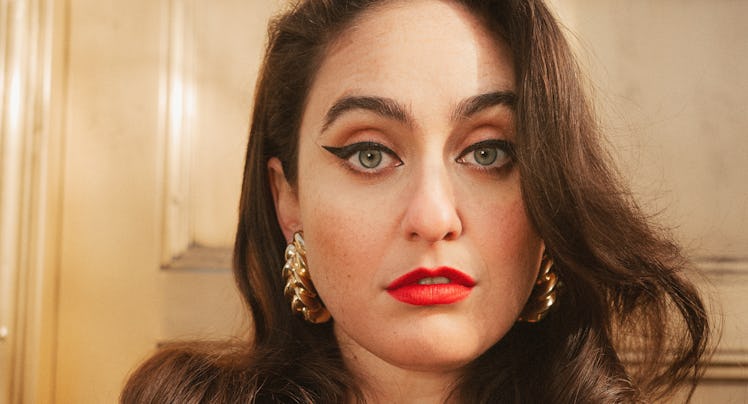 Comedian Catherine Cohen wearing a blazer, red lipstick and gold earrings.