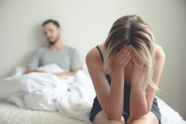 A frustrated wife sits on the edge of the bed with her head in her hands while her upset husband sit...