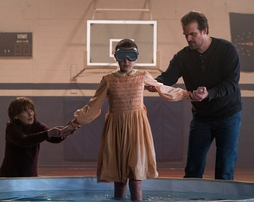 Joyce and Hopper help a blindfolded Eleven into a kiddie pool in a school gym.
