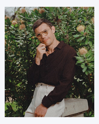 ryan o'connell wearing a black shirt and white pants in front of flowers