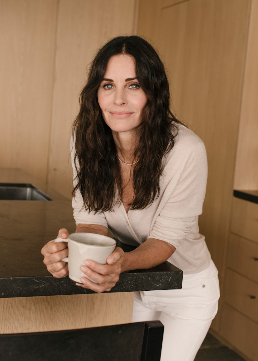 A portrait of Courtney wearing a white outfit, holding a mug in her hands, standing in a very clean ...