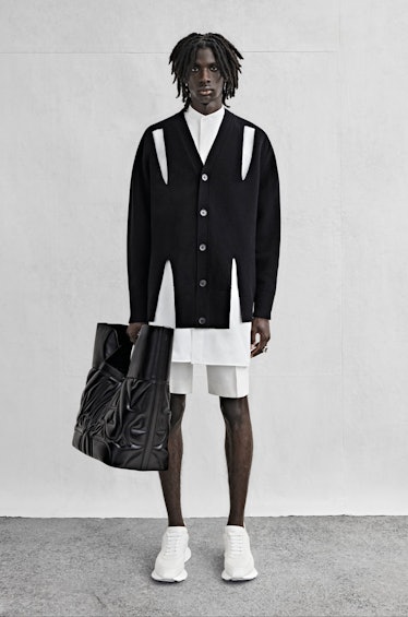 A model wearing an Alexander McQueen sweater, a white button down shirt and white shorts, carrying a...