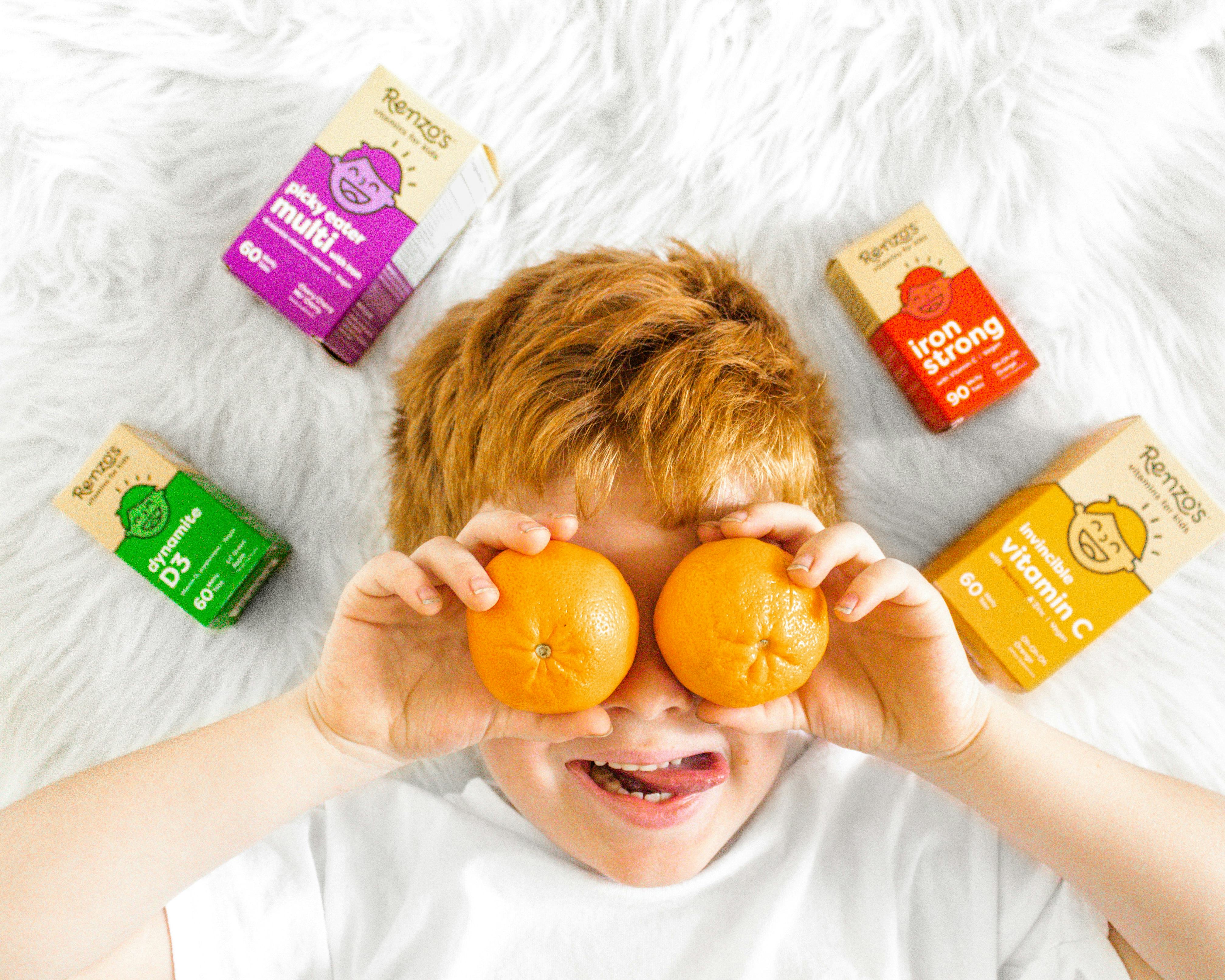 A picky eater boy placing two mandarins on his eyes with packages of vitamins spread on the floor ar...