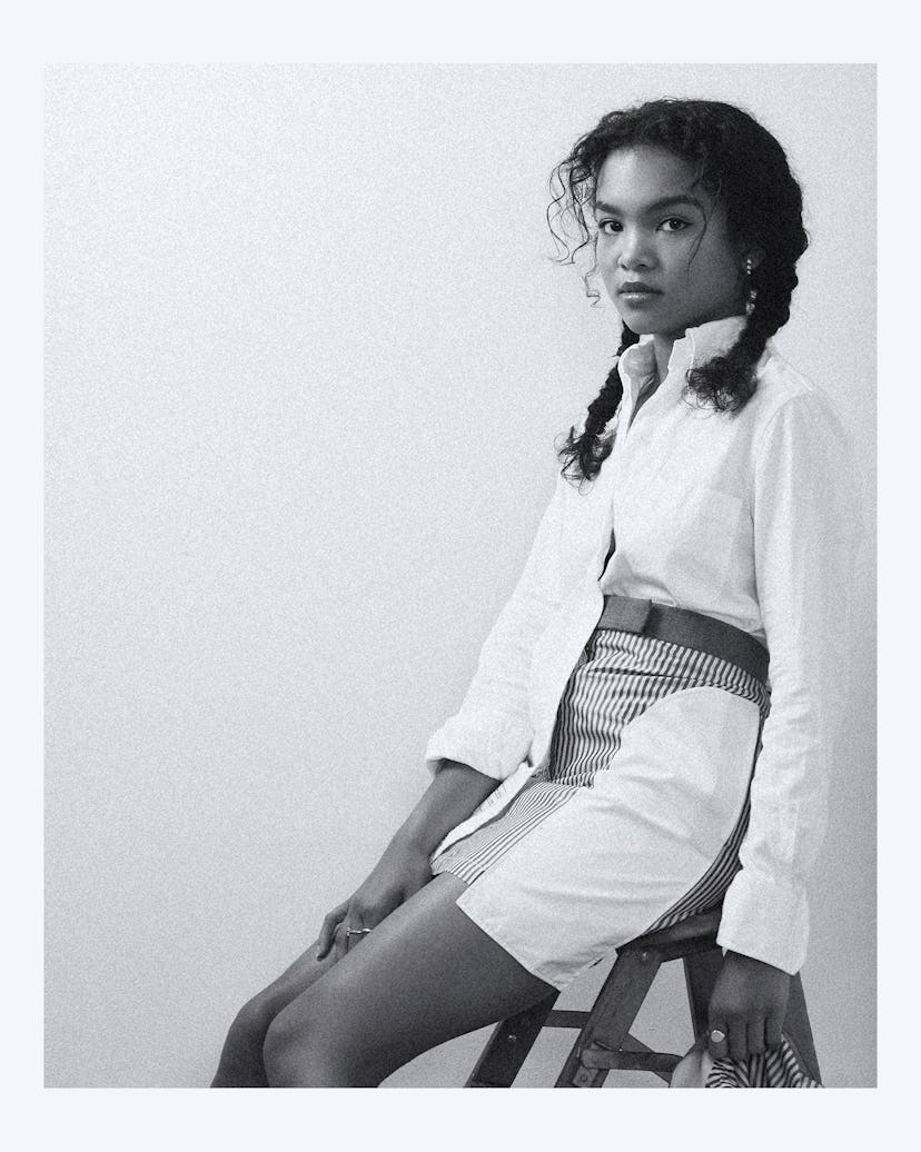 Mia Isaac sitting on a stool in a shirt and a skirt in black and white