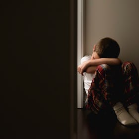 A depressed boy sits on the floor against a wall besides a doorframe that leads into the dark.