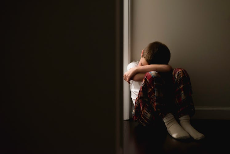 A depressed boy sits on the floor against a wall besides a doorframe that leads into the dark.