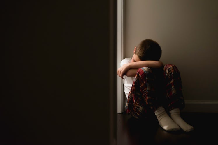 A depressed boy sits on the floor against a wall besides a doorframe that leads into the dark.