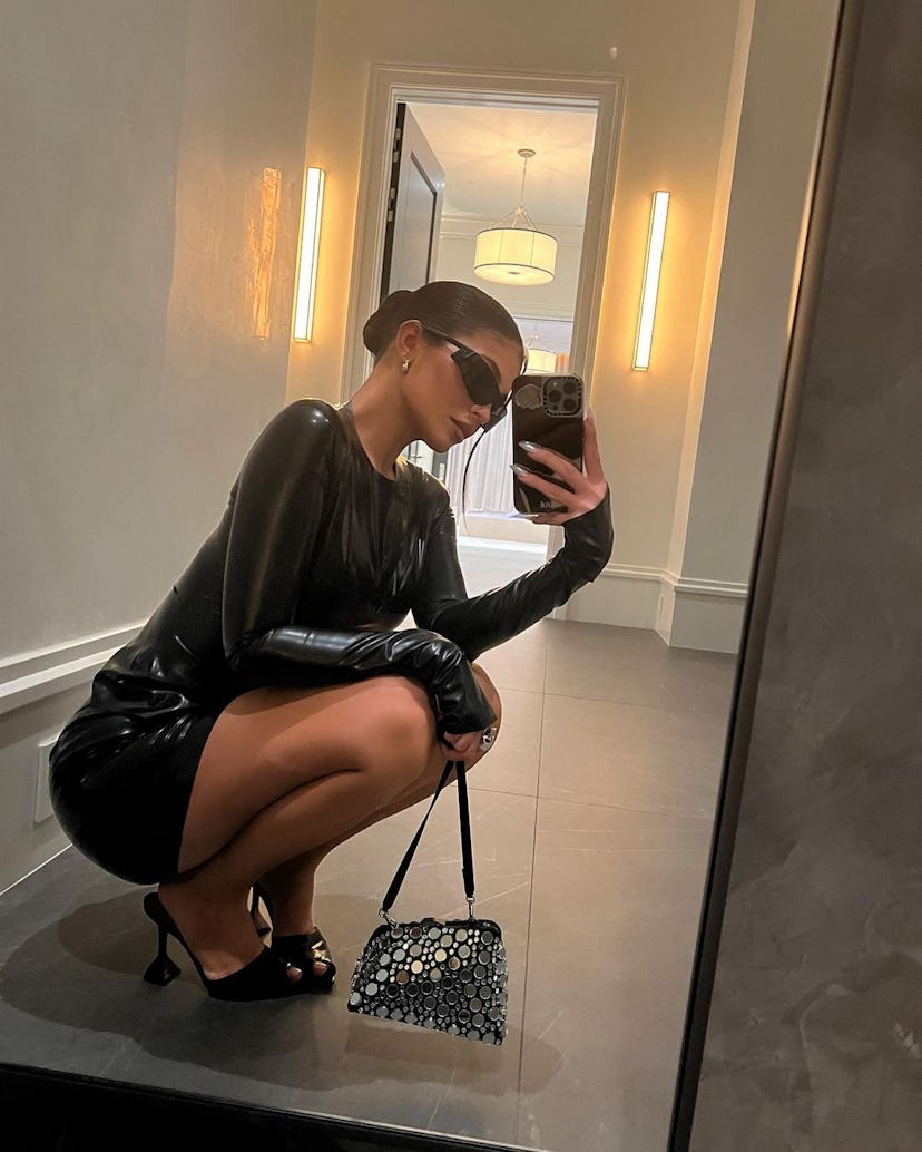 Kylie Jenner squatting in front of a mirror taking a selfie in a tight black latex mini dress with l...