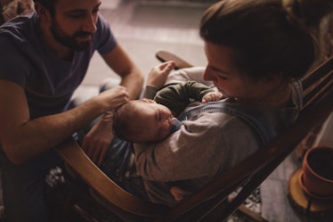 A mother in a rocking chair rocking her baby to sleep as dad kneels in front of them, smiling.