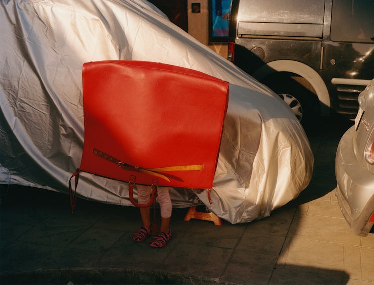 A little girl's legs protruding out of a giant, upside-down red bag, as she is in front of a car in ...