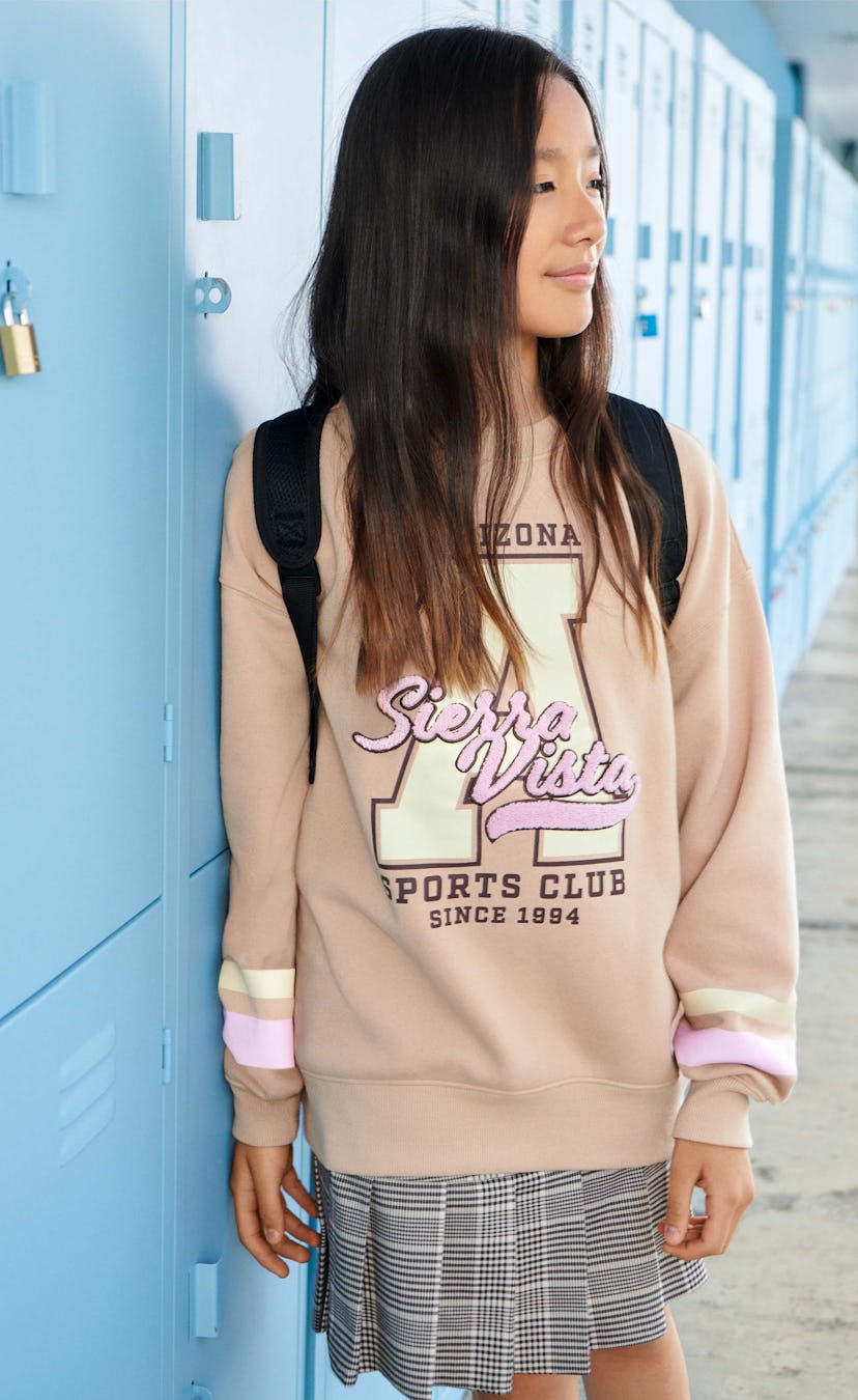 A girl leaned on blue school lockers, wearing a beige H&M oversized sweatshirt and a seamed pleated ...