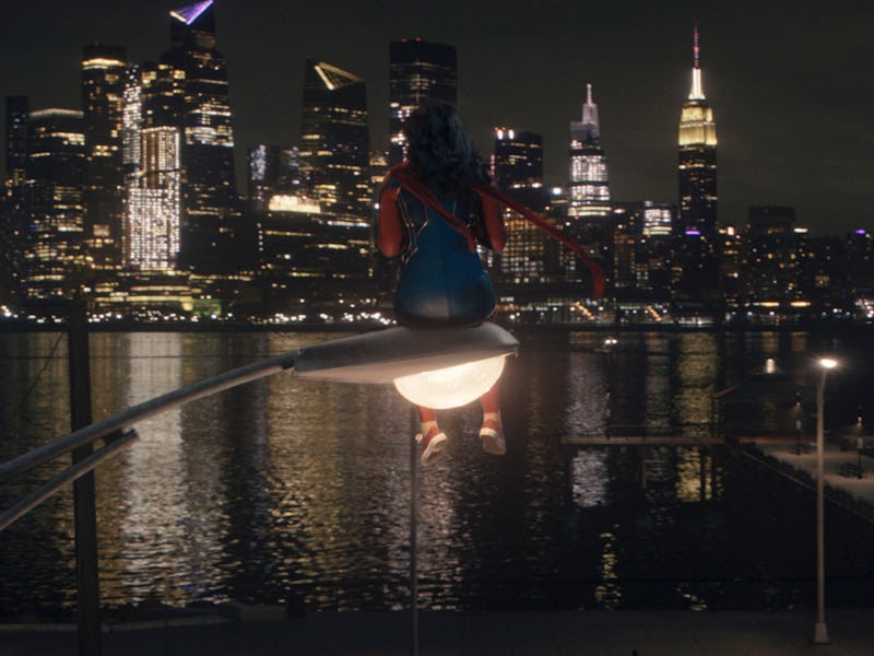 A scene with Ms. Marvel sitting on the top of a street lamp with the city lights in the background