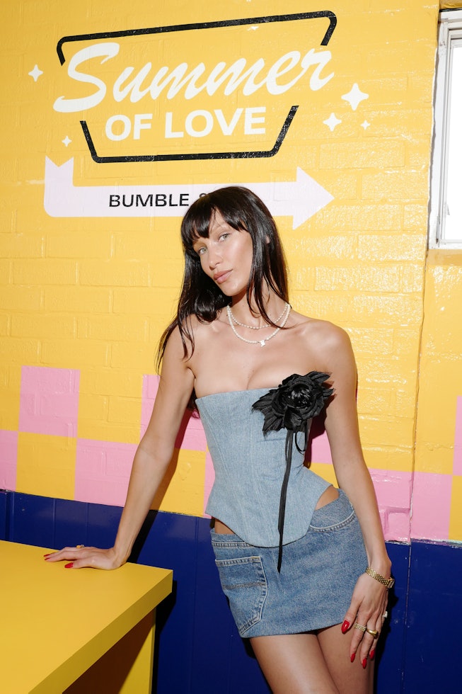 Bella Hadid in a denim short skirt and the same colored corset standing in front of a yellow wall th...