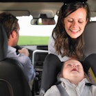 A dad drives a car while a mom smiles back at her baby in a rear-facing car seat.