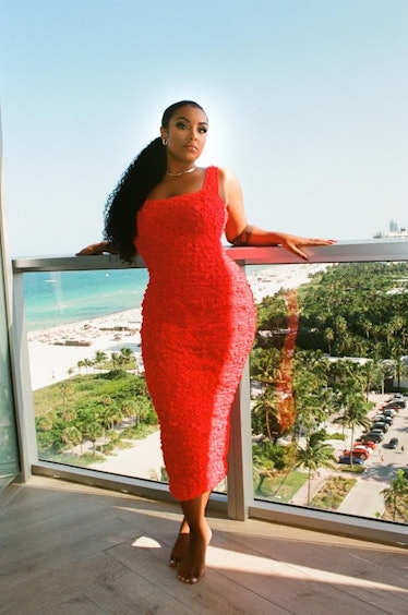 Aida Osman posing on the balcony with an ocean view behind her, wearing a tight, bright red dress.