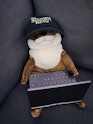 A plushy otter pictured wearing a baseball cap with the GPD Pocket 3 in its lap