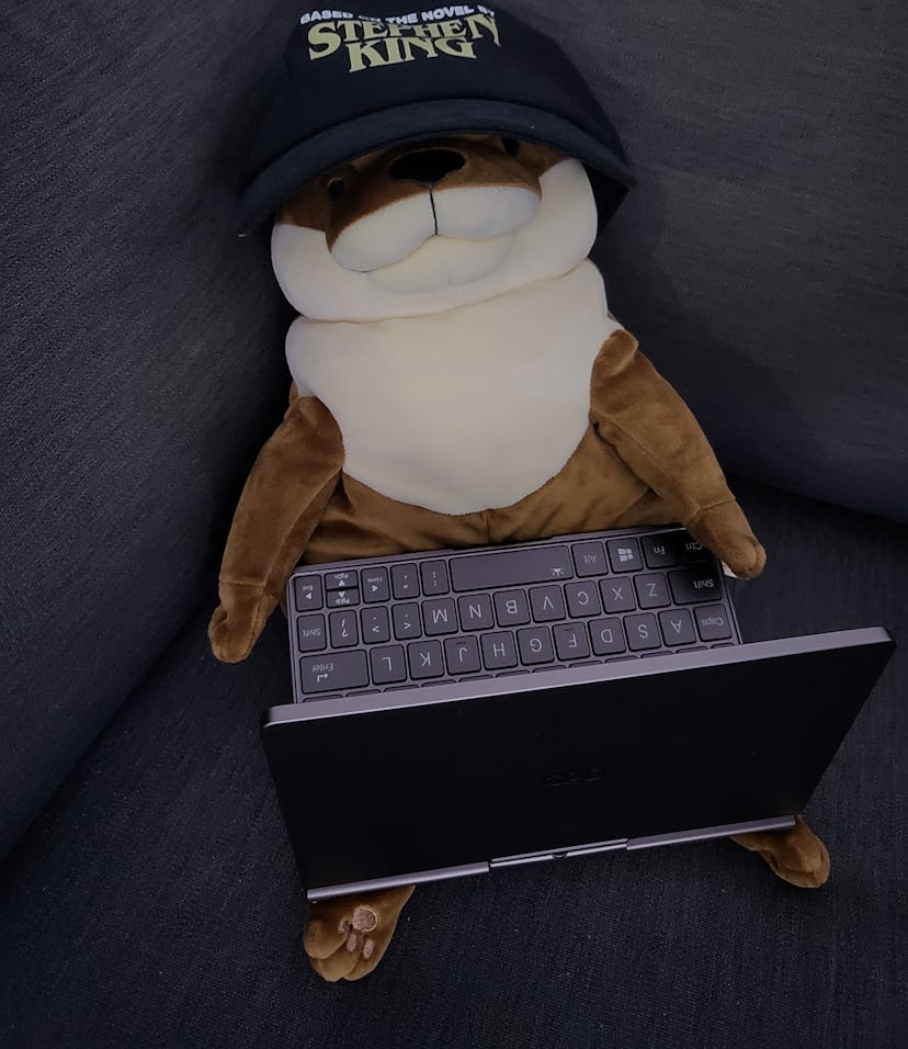 A plushy otter pictured wearing a baseball cap with the GPD Pocket 3 in its lap