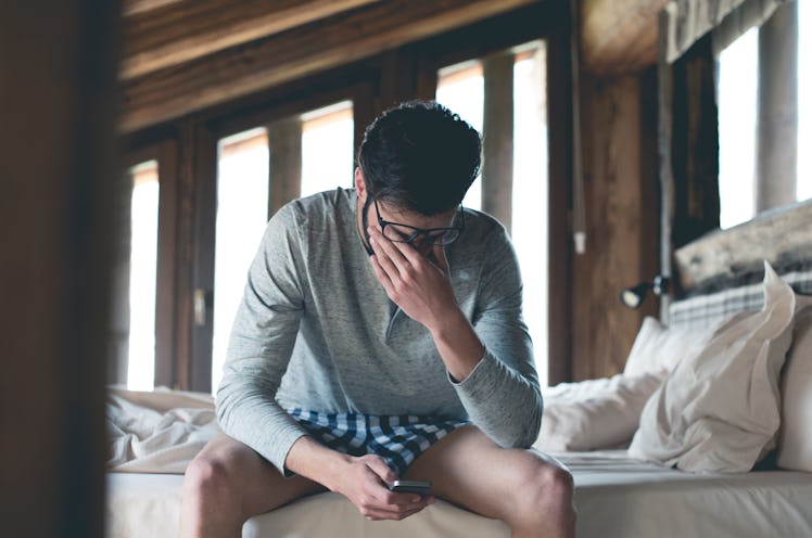 An unemployed man in his pajamas sits in bed, holding his face and phone and looking stressed.