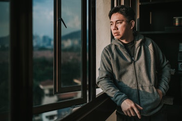 A depressed man in a hoodie looking out a window.