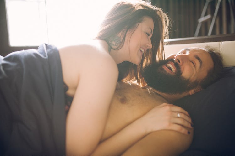 A man and woman laughing in bed, unclothed and covered by a blanket.