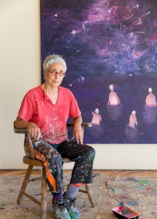 The artist Katherine Bradford, an older woman with cropped grey hair and glasses, wearing paint-spla...