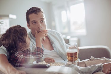A dad drinks beer as he and his son eat popcorn while watchin TV.