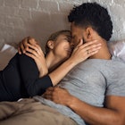 A short man lies with a woman in bed, smiling and kissing.