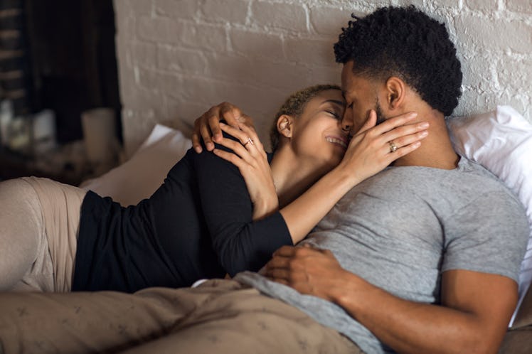 A short man lies with a woman in bed, smiling and kissing.