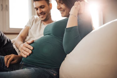 A dad-to-be puts his hand on his wife's baby bump as they sit on a couch.