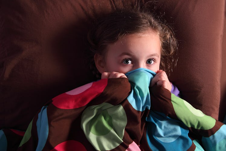 A scared child hiding her face under blankets in bed.