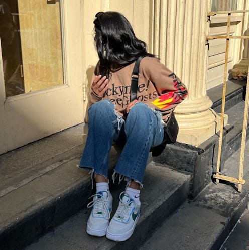 A woman in a brown sweater, blue denim jeans, and white footwear that looks fresh